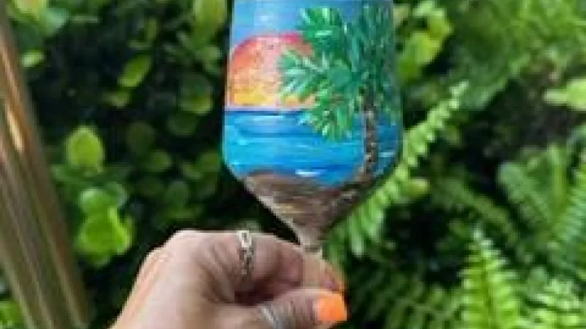 a hand with bright orange finger nails holding up a brightly painted wine glass with a beach scene on it
